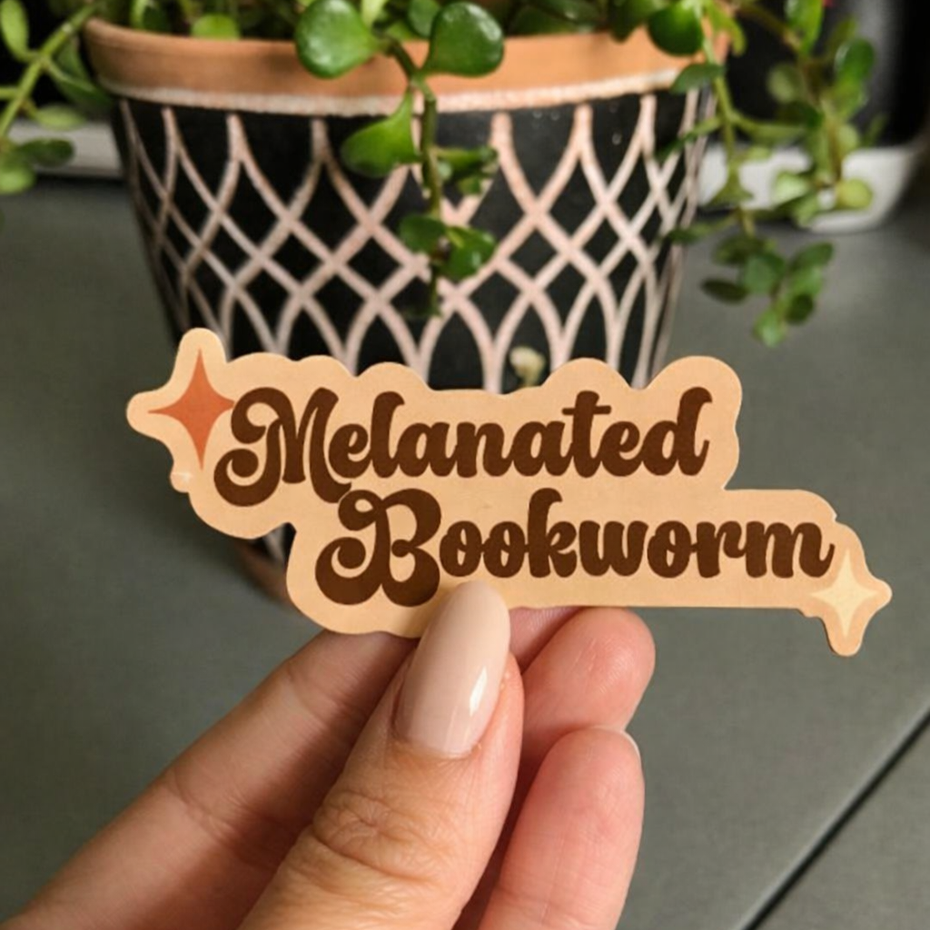 Hand holding a wooden sign with 'Melanated Bookworm' text in front of a potted plant.
