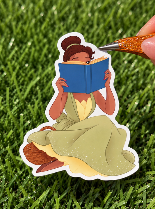 A die-cut sticker of a woman in a green dress reading a blue book, shown on a bed of green grass with tweezers holding the top edge of the sticker.